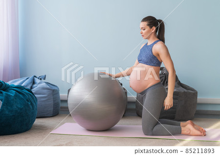 Pregnant european woman performs exercises with fitness ball. Expectant mother is doing yoga in the 3rd trimester. Girl in the classroom in preparation for childbirth. 85211893