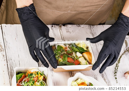Hands in black gloves are packing a business lunch in a cardboard box. Concept food delivery service. Delicious and healthy food at the workplace. Take away restaurant. Hands in black gloves are packing a business lunch in a cardboard box. Concept food delivery service. Delicious and healthy food at the workplace. Take away restaurant. 85214553