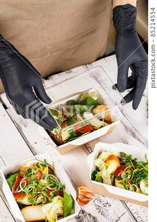 Hands in black gloves are packing a business lunch in a cardboard box. Concept food delivery service. Delicious and healthy food at the workplace. Take away restaurant. Vertical shot Hands in black gloves are packing a business lunch in a cardboard box. Concept food delivery service. Delicious and healthy food at the workplace. Take away restaurant. Vertical shot 85214554