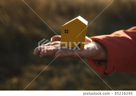 Close-up of female hand holding wooden white house model with autumn natural and a sunlight background Close-up of female hand holding wooden white house model with autumn natural and a sunlight background 85216295
