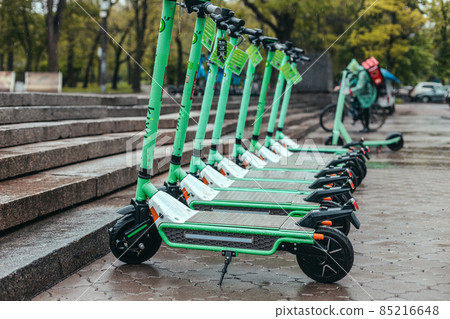 Row of green mint rental motorized kick scooters in the center of rainy city. Electric Urban transportation for every day. Row of green mint rental motorized kick scooters in the center of rainy city. Electric Urban transportation for every day. 85216648