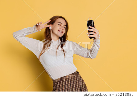 Cheerful young beautiful girl wearing warm white neck-polo in earphones using phone isolated on yellow color background. Emotions concept 85218083