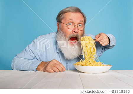 Shocked senior hairy gray-bearded man eating large portion of noodles, pasta isolated on blue studio background. 85218123