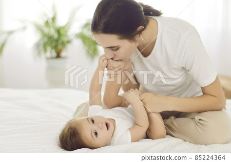 Caring mother play with little newborn daughter or son in bed at home. Loving young Caucasian mom caress comfort small baby kid child, enjoy family weekend together. Motherhood concept. 85224364