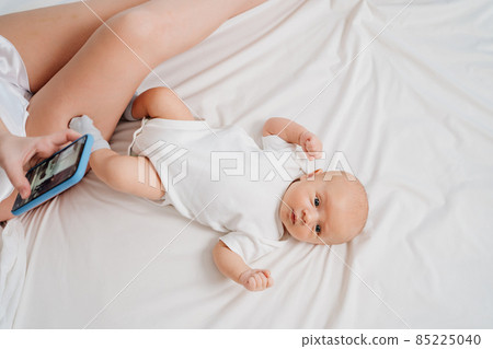 mom sits on the bed with a phone next to the baby. mom sits on the bed with a phone next to the baby. 85225040