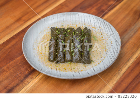 Delicious stuffed grape leaves. Traditional doom Mediterranean cuisine Dolma on white plate with fresh cilantro, on wooden background, close up. Delicious stuffed grape leaves. Traditional doom Mediterranean cuisine Dolma on white plate with fresh cilantro, on wooden background, close up. 85225320