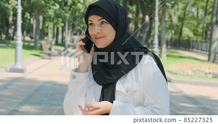 Happy arabian woman talking on smartphone on summer city park, close-up. Young female in black headscarf communicates by cellphone in sunny street outdoor. Smiling muslim girl speaking on mobile phone Happy arabian woman talking on smartphone on summer city park, close-up. Young female in black headscarf communicates by cellphone in sunny street outdoor. Smiling muslim girl speaking on mobile phone 85227325