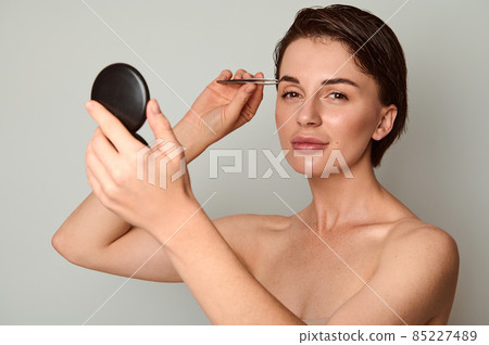 Charming wonderful brunette, young attractive woman with small cosmetic mirror and stainless steel tweezers, plucking her eyebrows, looking at camera, posing on light gray background with copy space 85227489