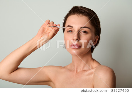 Beautiful dark haired Caucasian woman holding steel tweezers and removing hair from eyebrows posing against light gray background with copy ad space. Beauty treatment, grooming, face and skin care Beautiful dark haired Caucasian woman holding steel tweezers and removing hair from eyebrows posing against light gray background with copy ad space. Beauty treatment, grooming, face and skin care 85227490