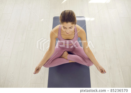 Healthy spirit in healthy body. Happy calm serene beautiful Caucasian woman sitting in easy cross legged lotus pose on yoga mat in gym, doing meditation, enjoying moment of zen, high angle, from above 85236570