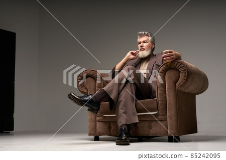 Full length shot of successful mature businessman with beard wearing elegant suit looking away, sitting with a glass of whisky in armchair over light gray background Full length shot of successful mature businessman with beard wearing elegant suit looking away, sitting with a glass of whisky in armchair over light gray background 85242095
