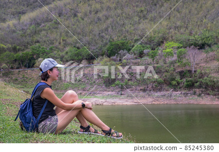 Side of a young asia woman sitting and enjoying peaceful moment of beautiful view at lake shore with mountains range in background. Side of a young asia woman sitting and enjoying peaceful moment of beautiful view at lake shore with mountains range in background. 85245380