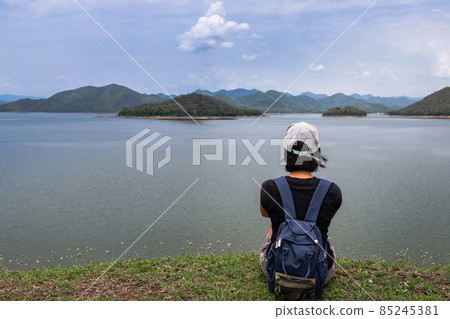 Back view of a young asia woman sitting and enjoying peaceful moment of beautiful view at lake shore with mountains range in background. Back view of a young asia woman sitting and enjoying peaceful moment of beautiful view at lake shore with mountains range in background. 85245381