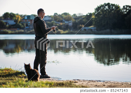Fisherman with spinning rod and cat near him on nature background. Angler man with fishing spinning or casting rod by the river. Fisherman with rod, spinning reel on the river bank 85247143