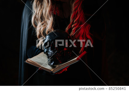 Witch in black costume with black magic book. Woman holds skull in hands. Death, spiritual rituals 85250841