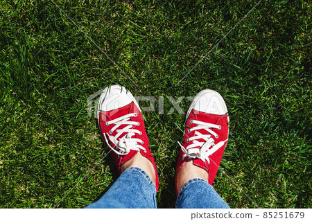 Legs in old red sneakers on green grass. View from above. The concept of youth, spring and freedom Legs in old red sneakers on green grass. View from above. The concept of youth, spring and freedom 85251679