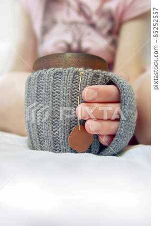 Cozy morning in bed with glanny mug of coffee, women's hands hold a mug in knitted cover. Close-up. Cozy morning in bed with glanny mug of coffee, women's hands hold a mug in knitted cover. Close-up. 85252557