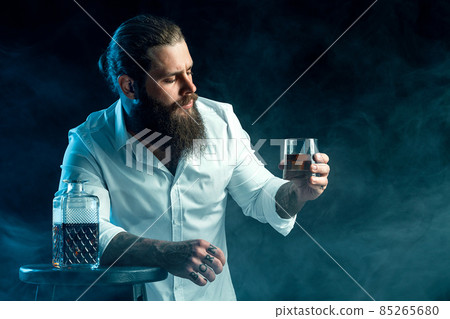bearded guy holds glass of whiskey, blows smoke on background, dressed in white shirt. Studio shot bearded guy holds glass of whiskey, blows smoke on background, dressed in white shirt. Studio shot 85265680