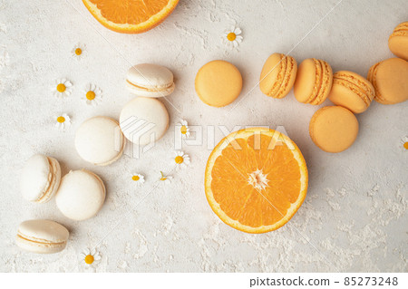 Orange and Vanilla French Macaroons with orange fruit filling on a creamy white clay background, decorated with small chamomiles and fresh sliced oranges 85273248