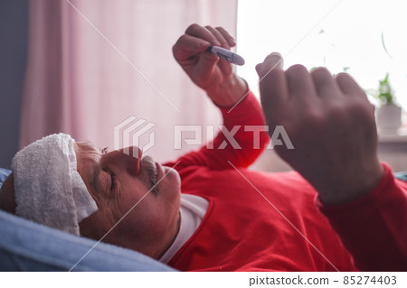 A sick male checking thermometer lying with wet towel on his forehead A sick male checking thermometer lying with wet towel on his forehead 85274403