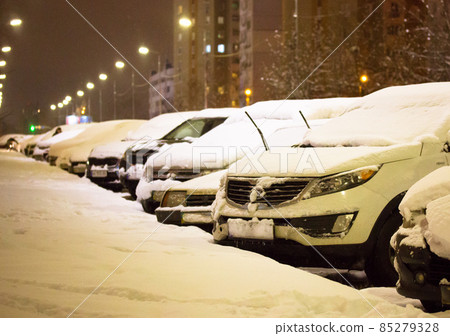 Many snow-covered parked cars buried in heavy snow parked along a street going into a distance in the evening, at night. City street in residential area after a heavy snowfall blizzard in winter. Many snow-covered parked cars buried in heavy snow parked along a street going into a distance in the evening, at night. City street in residential area after a heavy snowfall blizzard in winter. 85279328