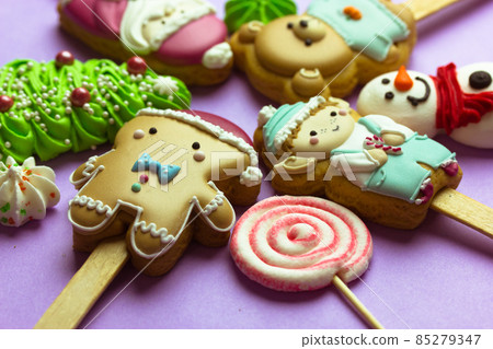Cookies on sticks in a shape of Santa Claus, elf character, Gingerbread man, snowman, bear, New Year 2022 tree, lollipop on purple background top view. Cute treats, sweets, candies for Christmas fun. Cookies on sticks in a shape of Santa Claus, elf character, Gingerbread man, snowman, bear, New Year 2022 tree, lollipop on purple background top view. Cute treats, sweets, candies for Christmas fun. 85279347