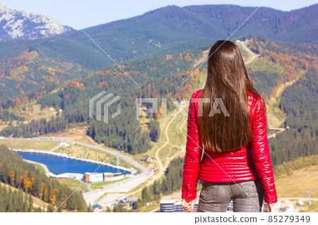 Beautiful cute slender girl in red down jacket, gray jeans staying on a top of a hill enjoying nature. Mountains, pond or river view in autumn sunny day. Young woman traveler back view, faceless trend Beautiful cute slender girl in red down jacket, gray jeans staying on a top of a hill enjoying nature. Mountains, pond or river view in autumn sunny day. Young woman traveler back view, faceless trend 85279349