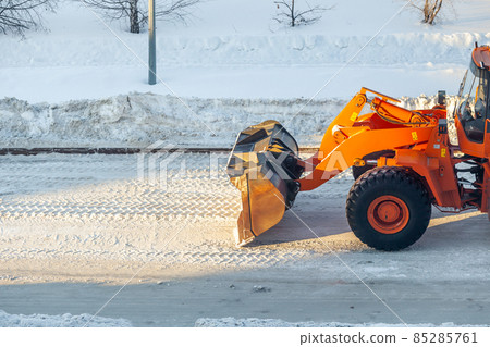 Big orange tractor cleans up snow from the road and loads it into the truck. Cleaning and cleaning of roads in the city from snow in winter 85285761