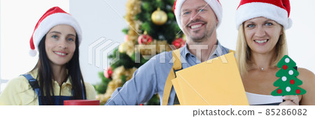 Group of couriers in santa claus hats standing near christmas tree and holding many gift boxes 85286082
