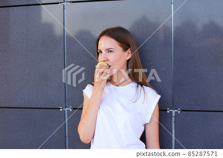 Young beautiful girl holds in hand and bites small green apple against background of gray wall. Young beautiful girl holds in hand and bites small green apple against background of gray wall. 85287571