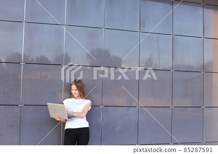 Beautiful young girl entrepreneur holds laptop and works, solves problems, and stands on background wall of business center. 85287591