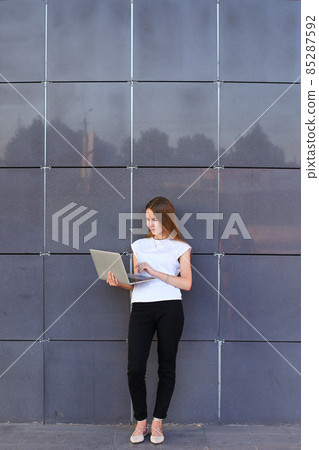 Perspective young girl entrepreneur holds laptop and works, solves problems, and stands on background wall of business center. Perspective young girl entrepreneur holds laptop and works, solves problems, and stands on background wall of business center. 85287592