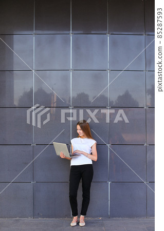 Perspective young girl entrepreneur holds laptop and works, solves problems, and stands on background wall of business center. Perspective young girl entrepreneur holds laptop and works, solves problems, and stands on background wall of business center. 85287593