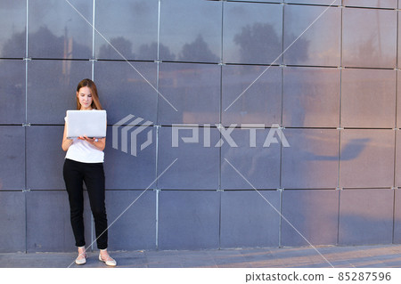 Perspective young girl entrepreneur holds laptop and works, solves problems, and stands on background wall of business center. 85287596