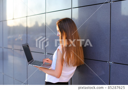 Attractive successful woman entrepreneur keeps in hands computer and works, typesets text, resolves issues on background of wall of business center. Attractive successful woman entrepreneur keeps in hands computer and works, typesets text, resolves issues on background of wall of business center. 85287625