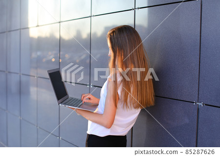 Attractive successful woman entrepreneur keeps in hands computer and works, typesets text, resolves issues on background of wall of business center. Attractive successful woman entrepreneur keeps in hands computer and works, typesets text, resolves issues on background of wall of business center. 85287626