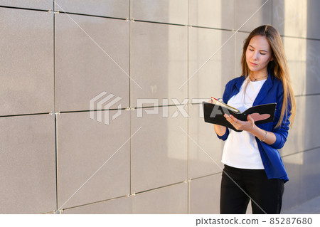 Young adult female girl student holds in hands diary, notebook, Students' work, homework and writes pen against backdrop of business center. Young adult female girl student holds in hands diary, notebook, Students' work, homework and writes pen against backdrop of business center. 85287680