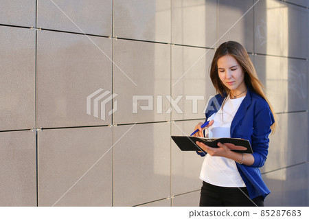 Young adult female girl student holds in hands diary, notebook, Students' work, homework and writes pen against backdrop of business center. Young adult female girl student holds in hands diary, notebook, Students' work, homework and writes pen against backdrop of business center. 85287683