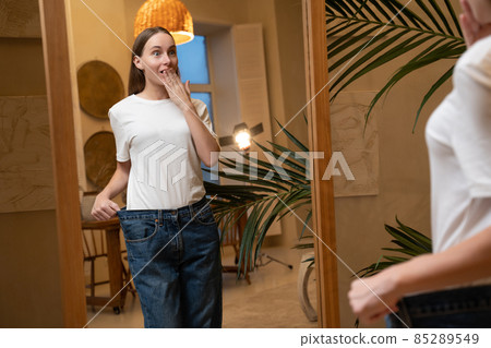 A woman who has lost a lot of weight due to a diet looks in the mirror and shows big jeans A woman who has lost a lot of weight due to a diet looks in the mirror and shows big jeans 85289549