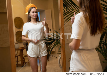 Slender woman standing in front of a mirror and measuring her waist after losing weight 85289551