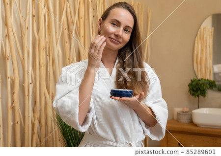 Smiling beautiful woman in a bathrobe puts a skin care cream on her face. A woman looks in the bathroom mirror. Morning healthy skin care Smiling beautiful woman in a bathrobe puts a skin care cream on her face. A woman looks in the bathroom mirror. Morning healthy skin care 85289601