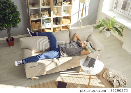 Relaxed guy sleeping on comfortable beige couch at home. Man taking break from work and napping on soft couch with laptop on coffee table beside in cosy modern living room. High angle. Rest concept 85290273