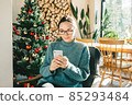 A young beautiful European woman uses her phone at home in a Christmas festive atmosphere. Concept greeting New Year 85293484