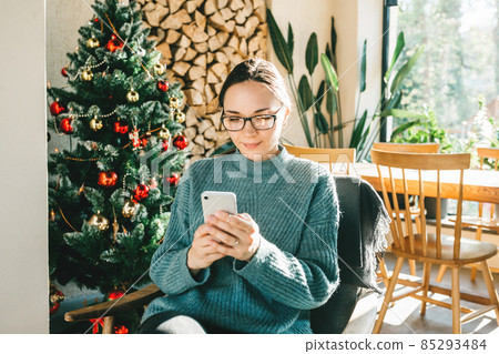 A young beautiful European woman uses her phone at home in a Christmas festive atmosphere. Concept greeting New Year 85293484