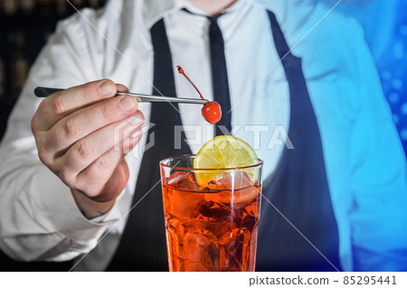 A professional bartender puts a cherry in a red chilled alcoholic cocktail with bar tweezers in glass at a nightclub counter 85295441