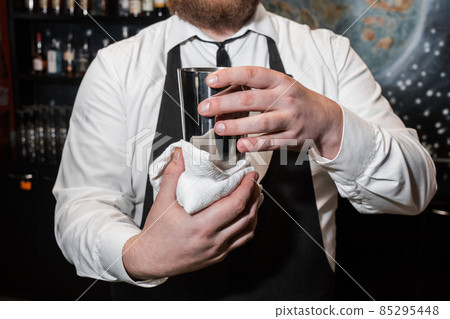 A professional bartender holds a metal shaker tool for mixing and making alcoholic cocktails and wipes it with a white cloth 85295448