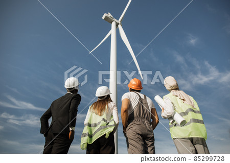 Multicultural partners in helmets looking on wind turbine Multicultural partners in helmets looking on wind turbine 85299728