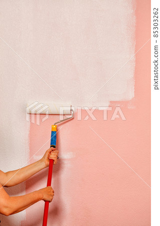 The painter's female hands hold a paint roller on a long telescopic rod against the background of a pink wall. The painter's female hands hold a paint roller on a long telescopic rod against the background of a pink wall. 85303262