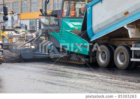 Construction of a new road, a truck pours hot asphalt into the bucket of an asphalt paver. Close-up. Construction of a new road, a truck pours hot asphalt into the bucket of an asphalt paver. Close-up. 85303263