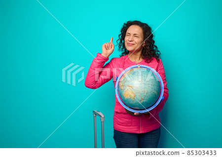 Beautiful woman in bright pink jacket pointing her finger up against blue background and posing with closed eyes, ready to point to travel destination on globe map. Travel, tourism concept. Copy space 85303433